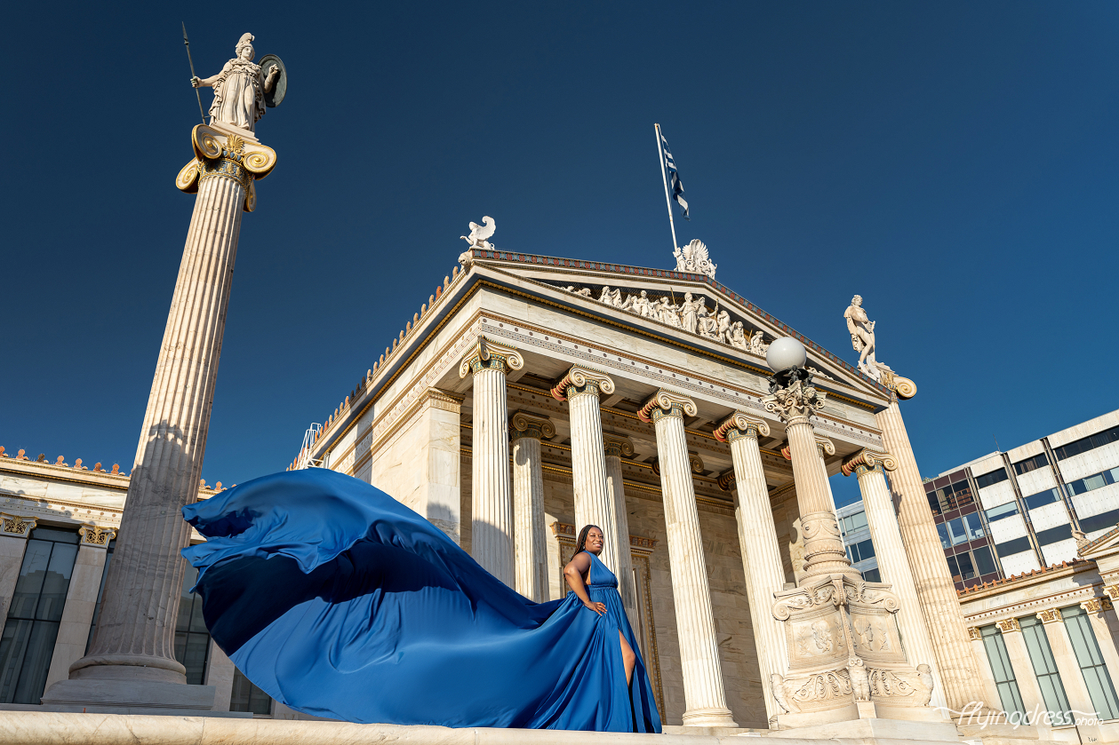 Woman in a royal blue satin flying dress posing on the steps of the Academy of Athens, with the dress sweeping dramatically against the neoclassical columns and the statue of Athena.